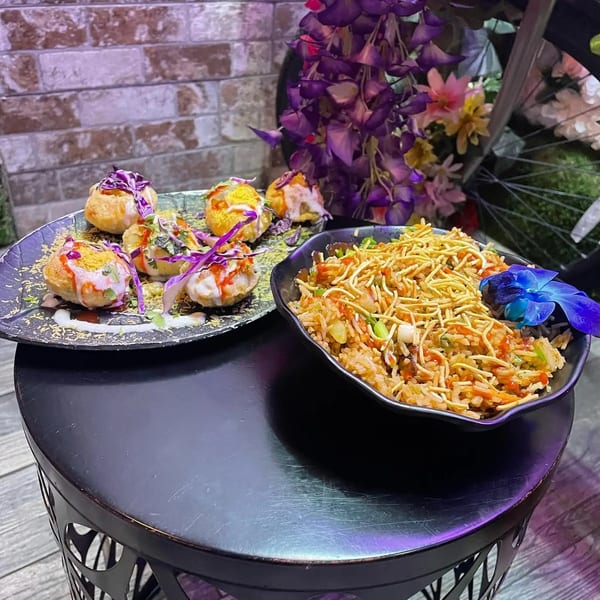 A black plate of pani puri and a black bowl of fried rice with noodles, on a black table.